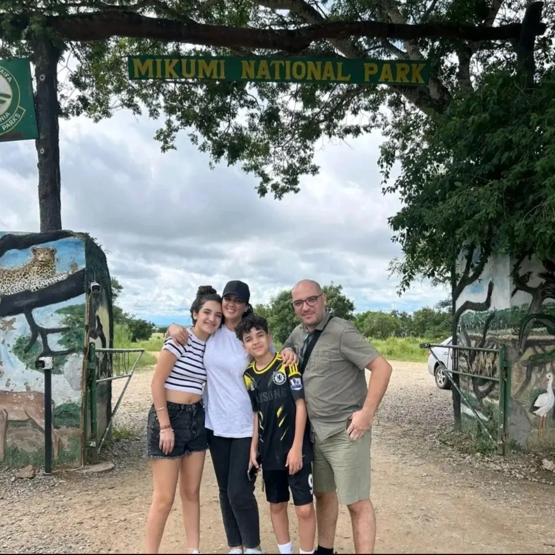 Family group photo standing before the Mikumi National Park main gate with the iconic welcome stone stele and green savanna trees in the background.